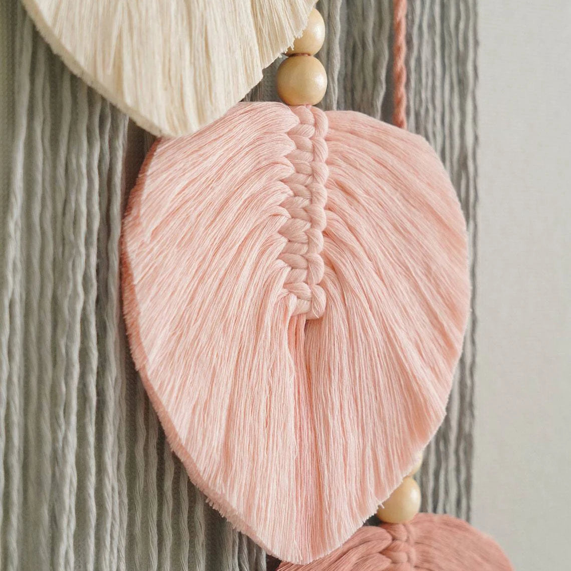 Decorative wall hanging with pink leaf-shaped tassels and wooden beads on a textured background